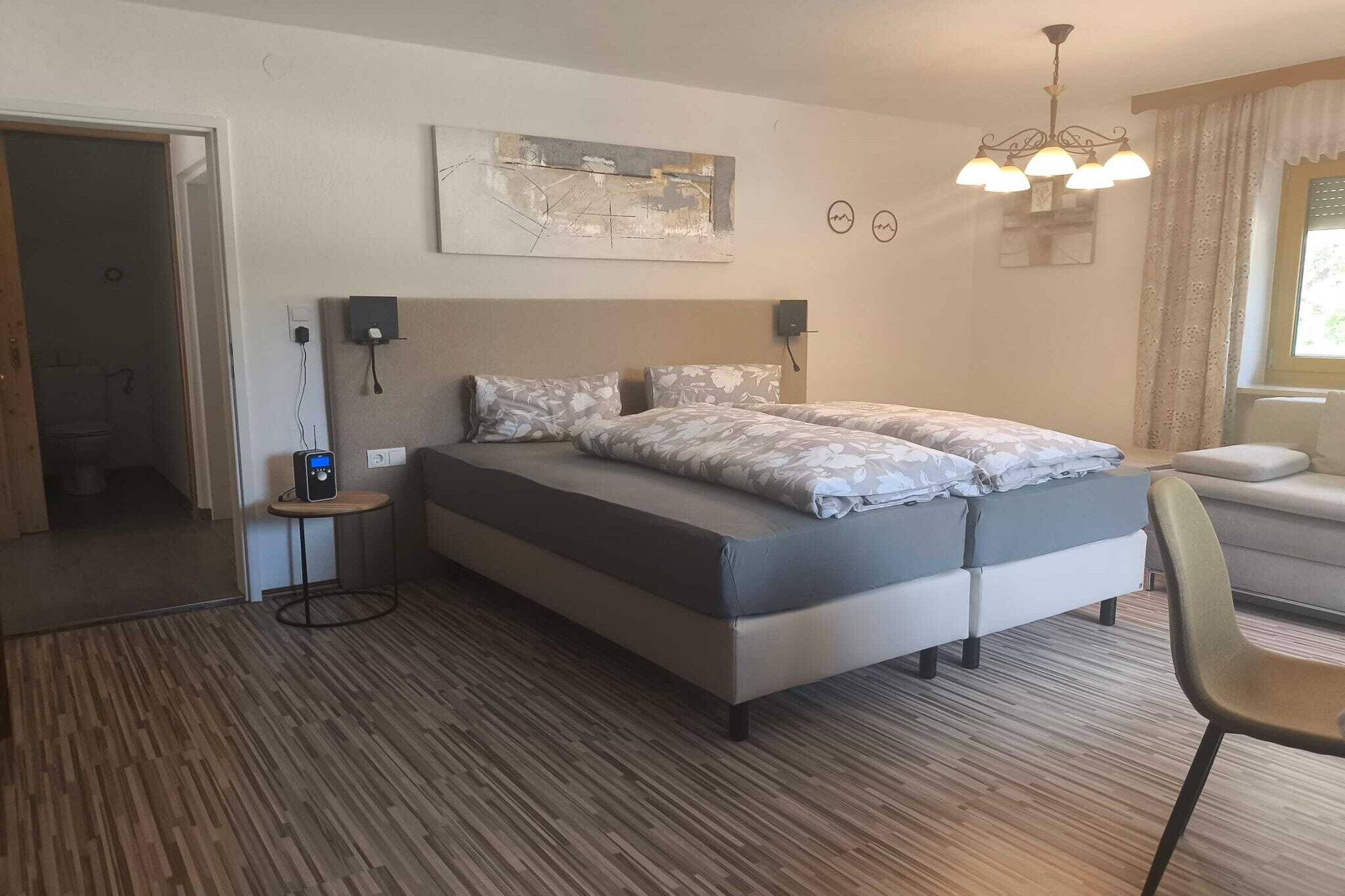 Une chambre d’hôtel avec un lit, une table de chevet avec une radio, une horloge murale et une fenêtre avec des rideaux.