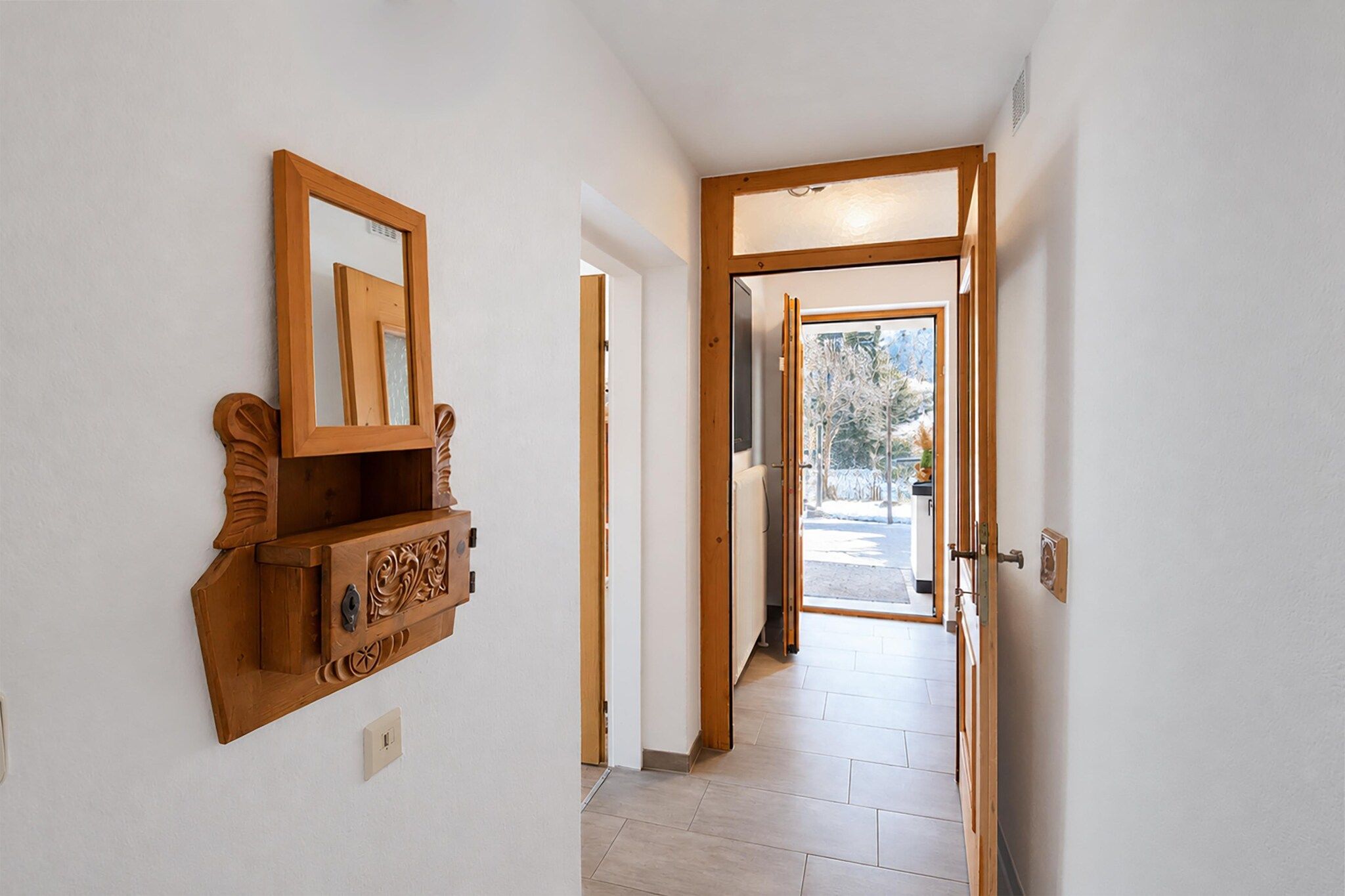 Un couloir avec une armoire en bois et un miroir, qui mène à une porte ouverte donnant sur un espace extérieur enneigé.