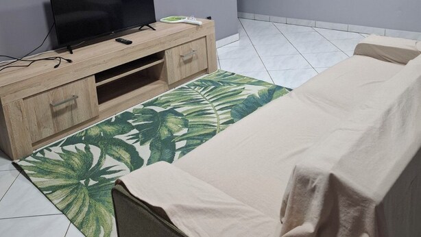 Una sala de estar con un televisor de pantalla plana sobre un mueble de madera, un tapete verde con estampado, un sofá beige y una pared con un cuadro floral.