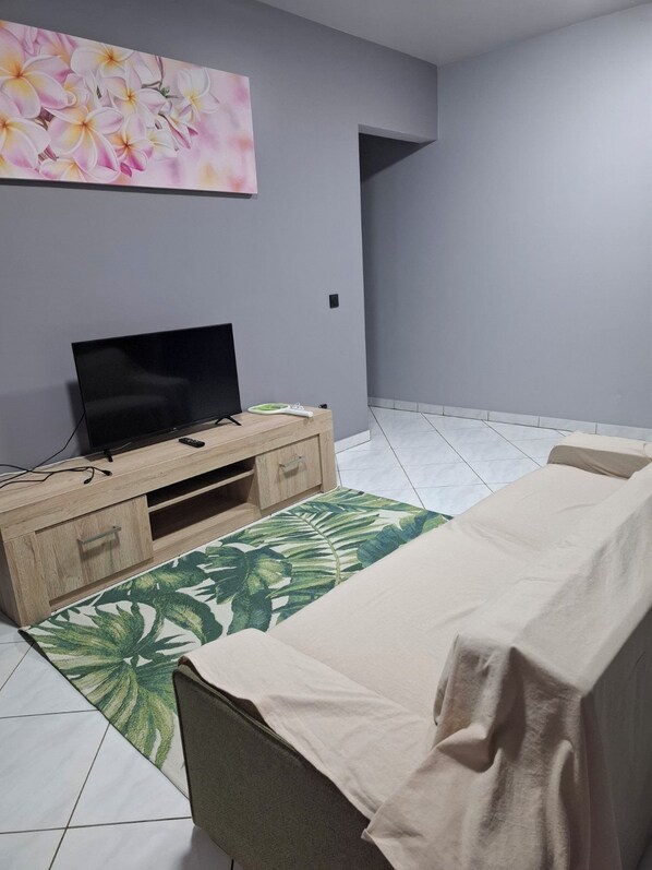 Un salon avec un téléviseur à écran plat posé sur un meuble en bois, un tapis à motifs verts, un canapé beige et un mur orné d’un tableau floral.