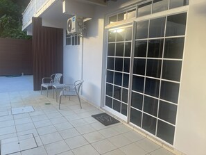 Un bâtiment moderne avec une façade élégante, une grande porte-fenêtre coulissante, un petit espace extérieur aménagé avec des chaises et un appareil de climatisation fixé au mur.