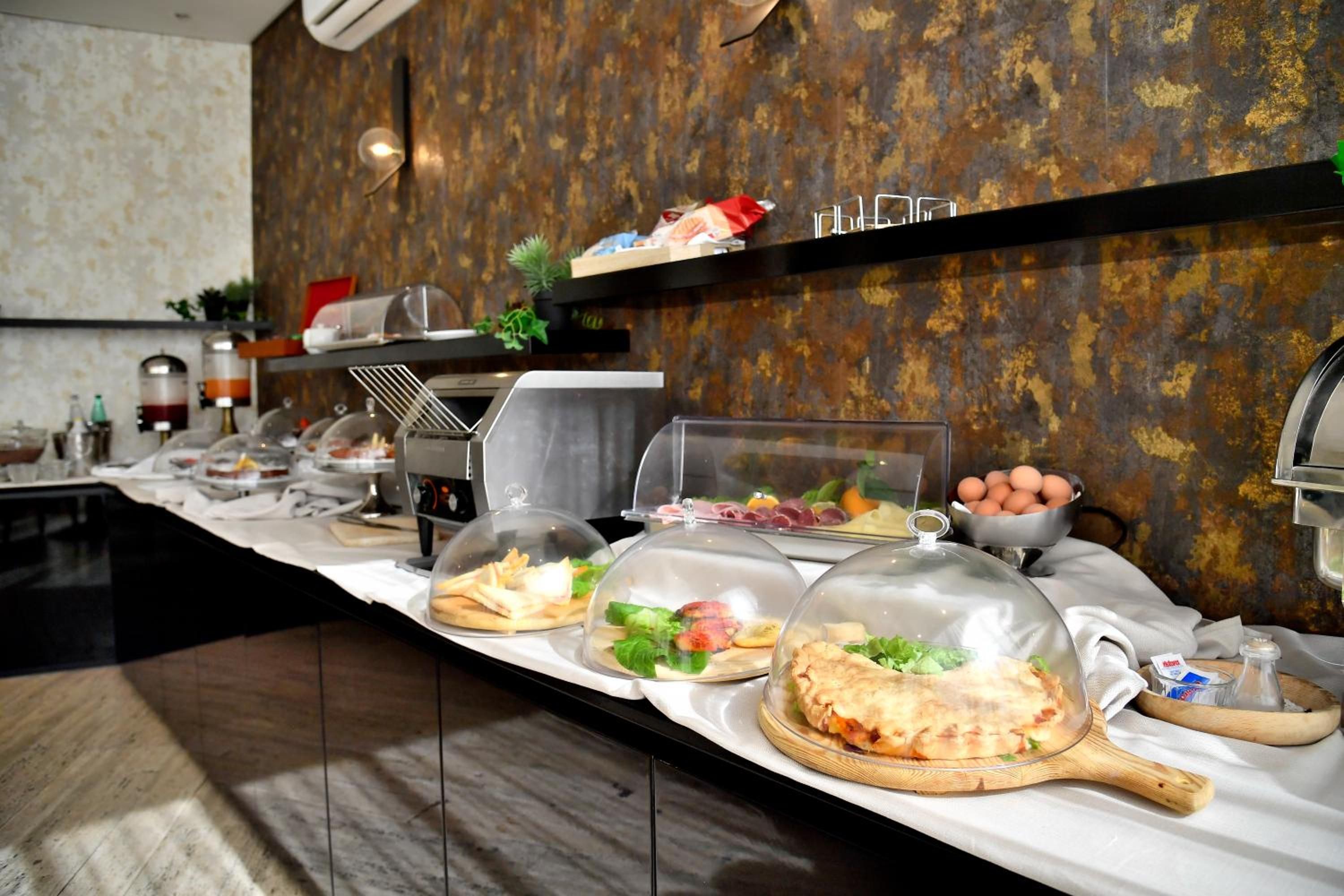 Ein Buffet mit verschiedenen Speisen unter Glasglocken, einer Kaffeemaschine und einer Wand mit strukturierter Oberfläche.
