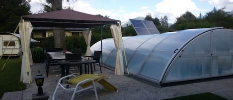 A greenhouse structure with a transparent roof, a covered seating area with a table and chairs, and a small lamp post.