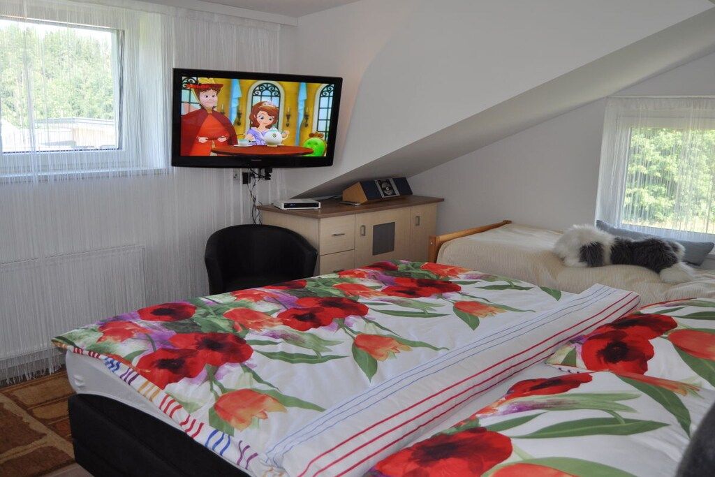 A bedroom with a bed, a television mounted on the wall, a window with curtains, and a small table with a picture frame.
