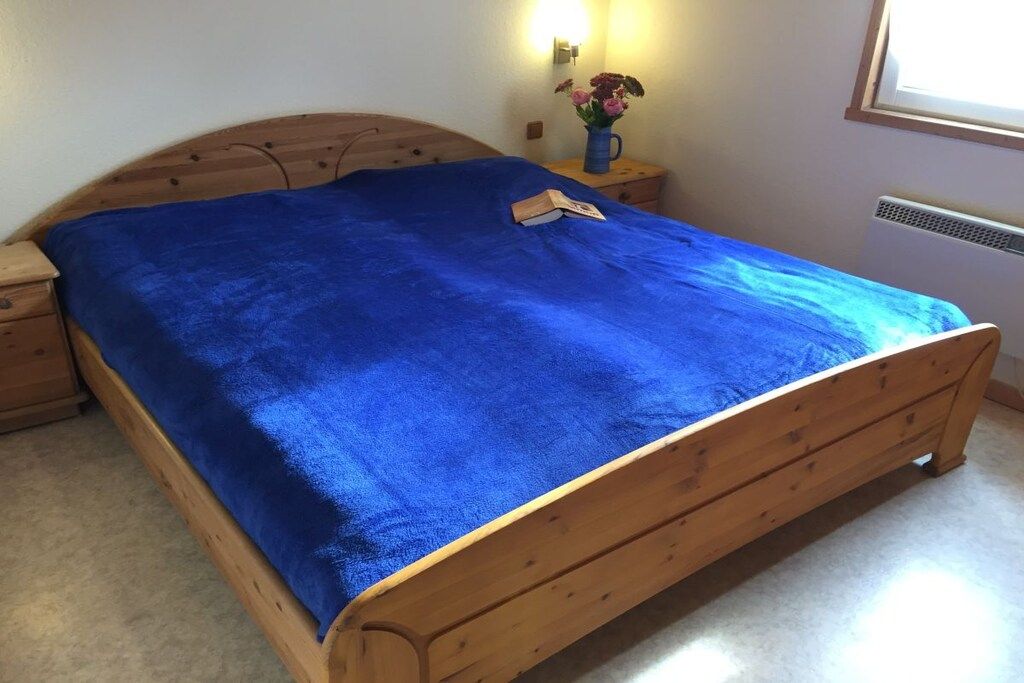 Ein hölzernes Bett mit einer blauen Decke, ein Nachttisch mit einer Lampe und ein Fenster mit Blick.