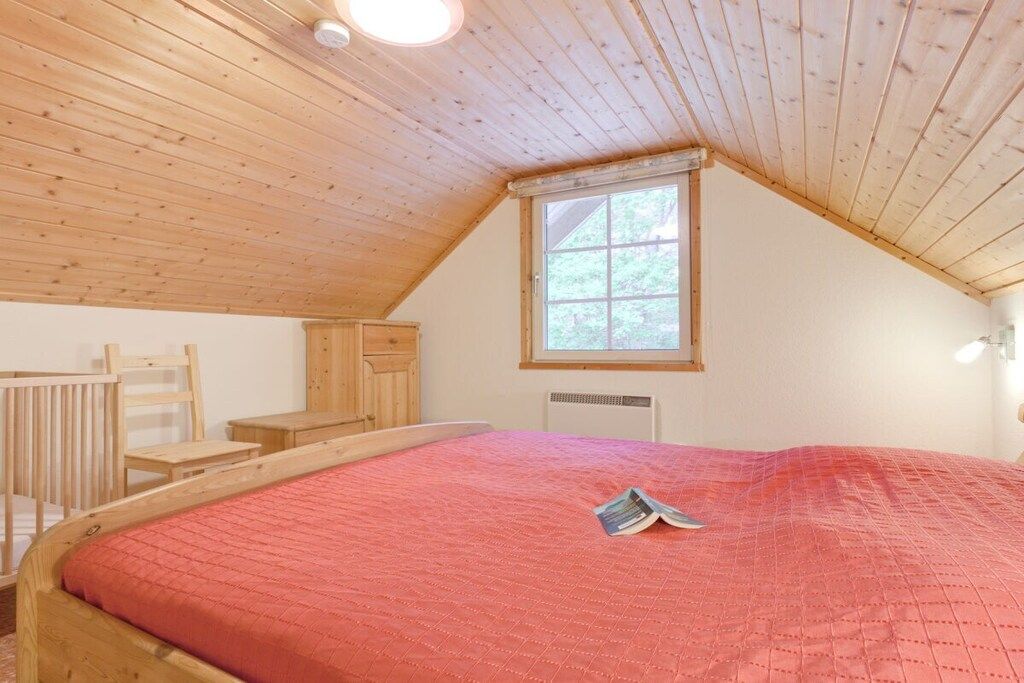Ein Schlafzimmer mit schrägen Wänden, einem Fenster, einem Bett mit roter Bettdecke, einem Holzstuhl und einem kleinen Tisch.