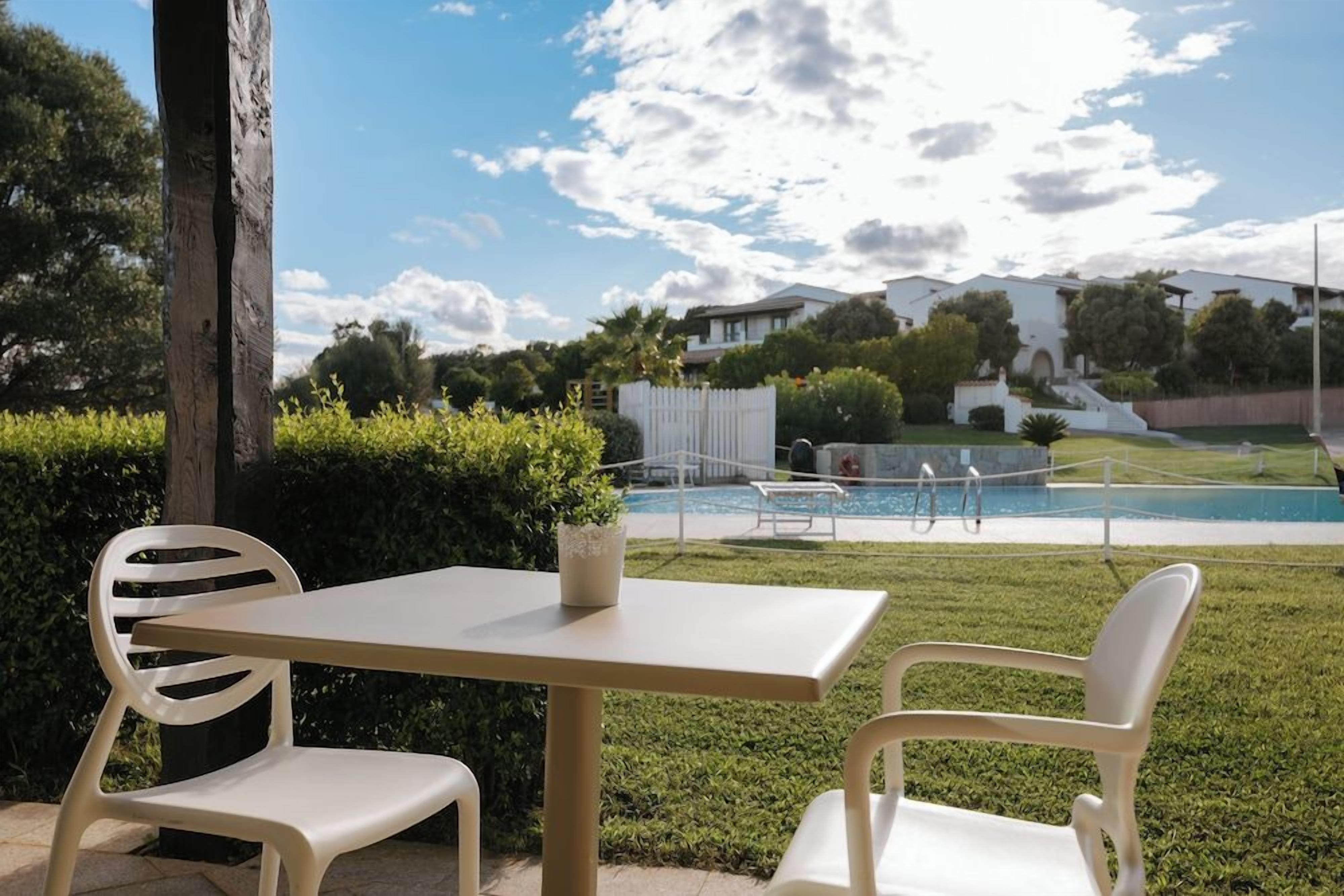 An outdoor seating area with a table and chairs, a potted plant, and a view of a swimming pool and houses in the background.
