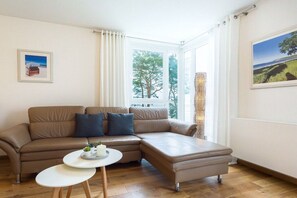 A modern living room with a brown leather sofa, a white coffee table, and a large window with a view of trees. - 6 (Seagull) in Villa Helene (Ostseebad Binz)