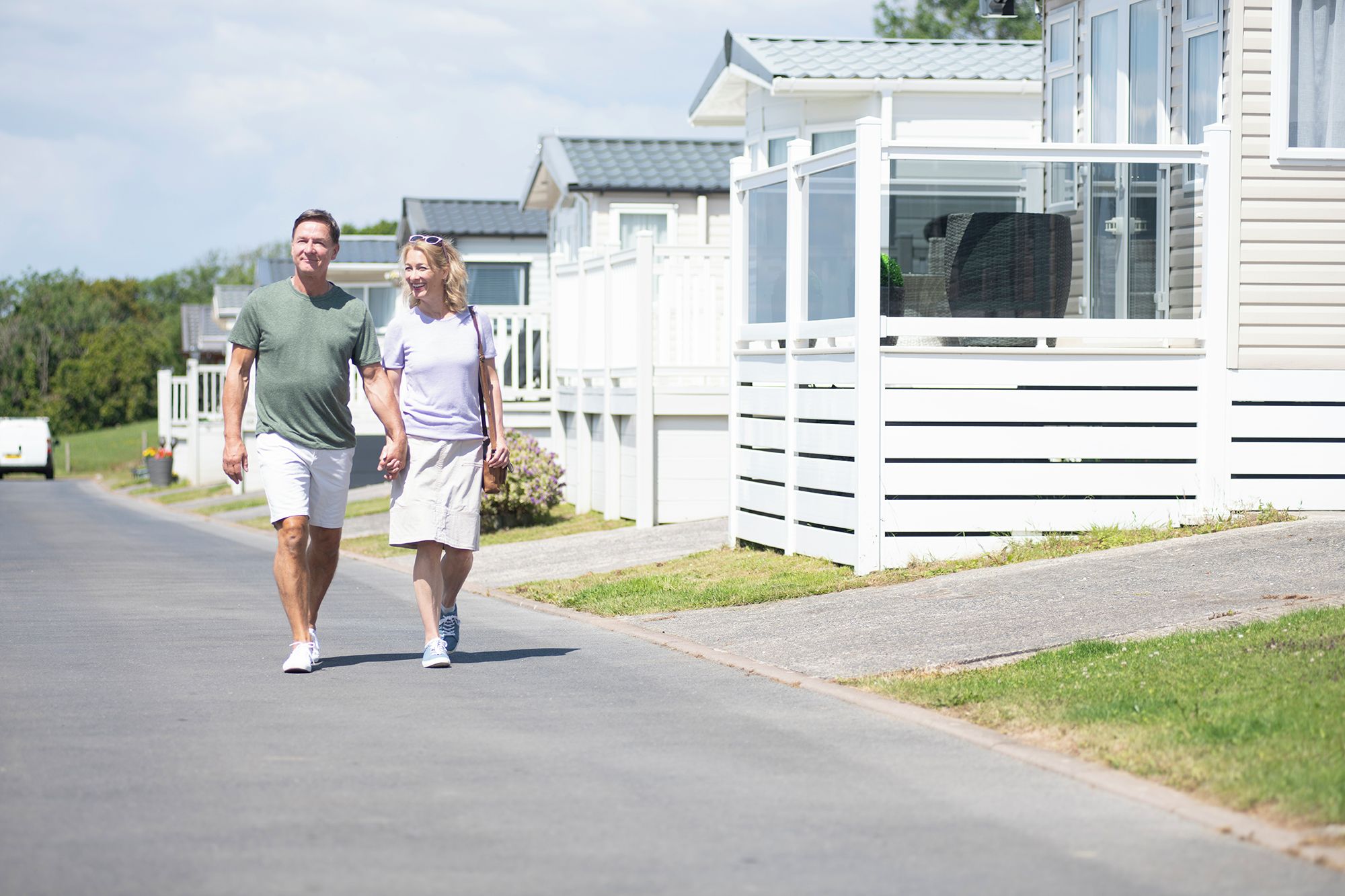 A couple walking on a paved road in a residential area with houses and a white fence.