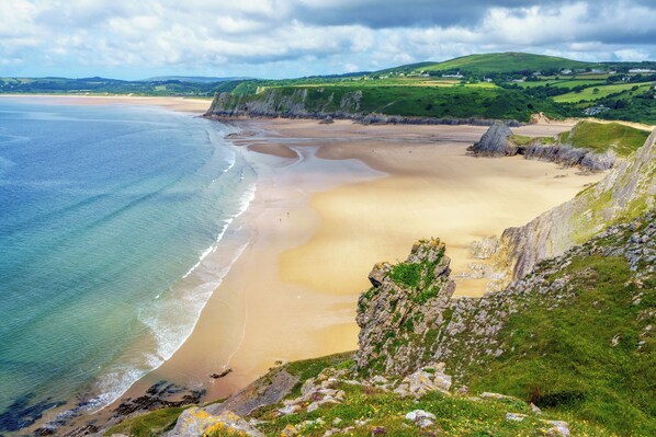 A coastal landscape with a sandy beach, cliffs, and rolling hills in the background. - Llanrhidian Holiday Park (Swansea)