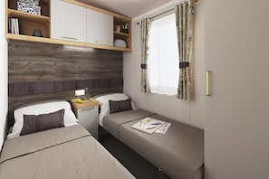 A compact bedroom with two beds, a bedside table, a shelf with books, and a window with curtains.
