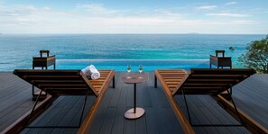 Terraza de madera con sillones de descanso y una mesa de vidrio, con vista al mar azul cristalino e una piscina infinita.