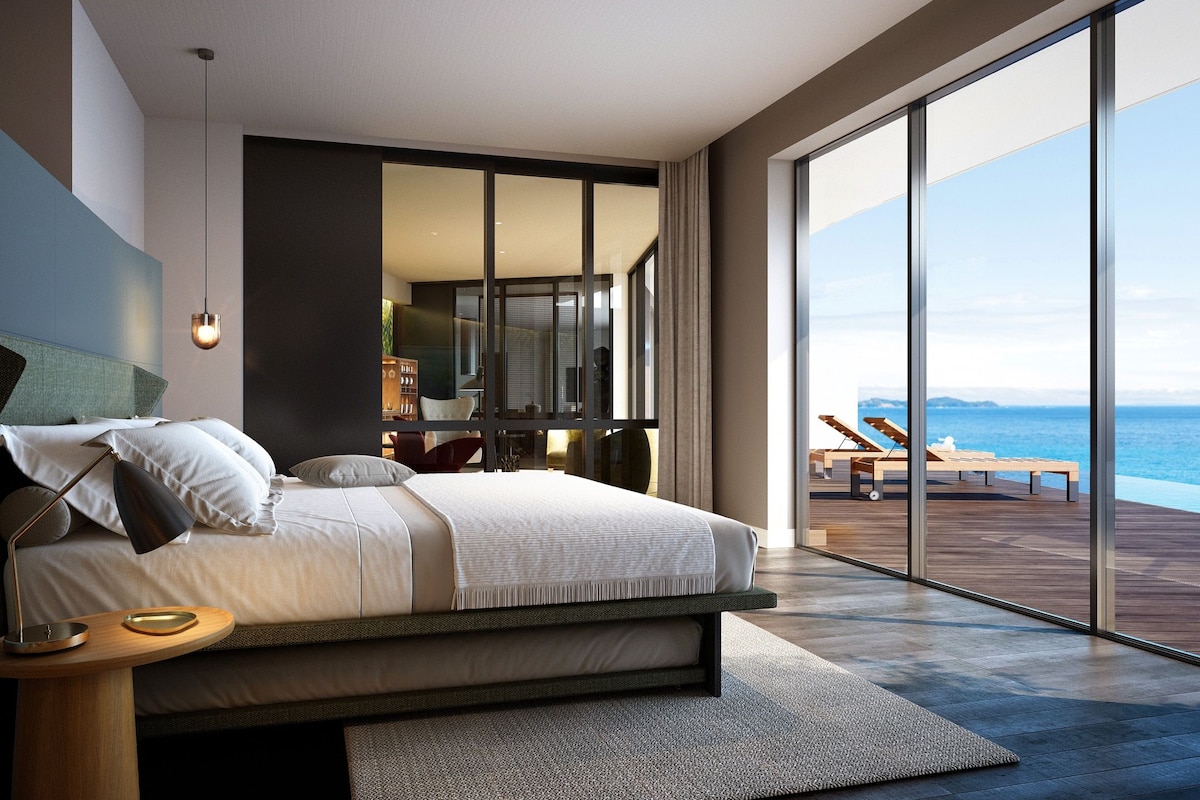 A modern bedroom with a large bed, a nightstand, and a view of the ocean through floor-to-ceiling windows.