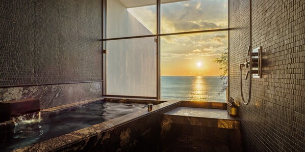 Un baño con bañera, ducha y un amplio ventanal con vista al mar al atardecer.
