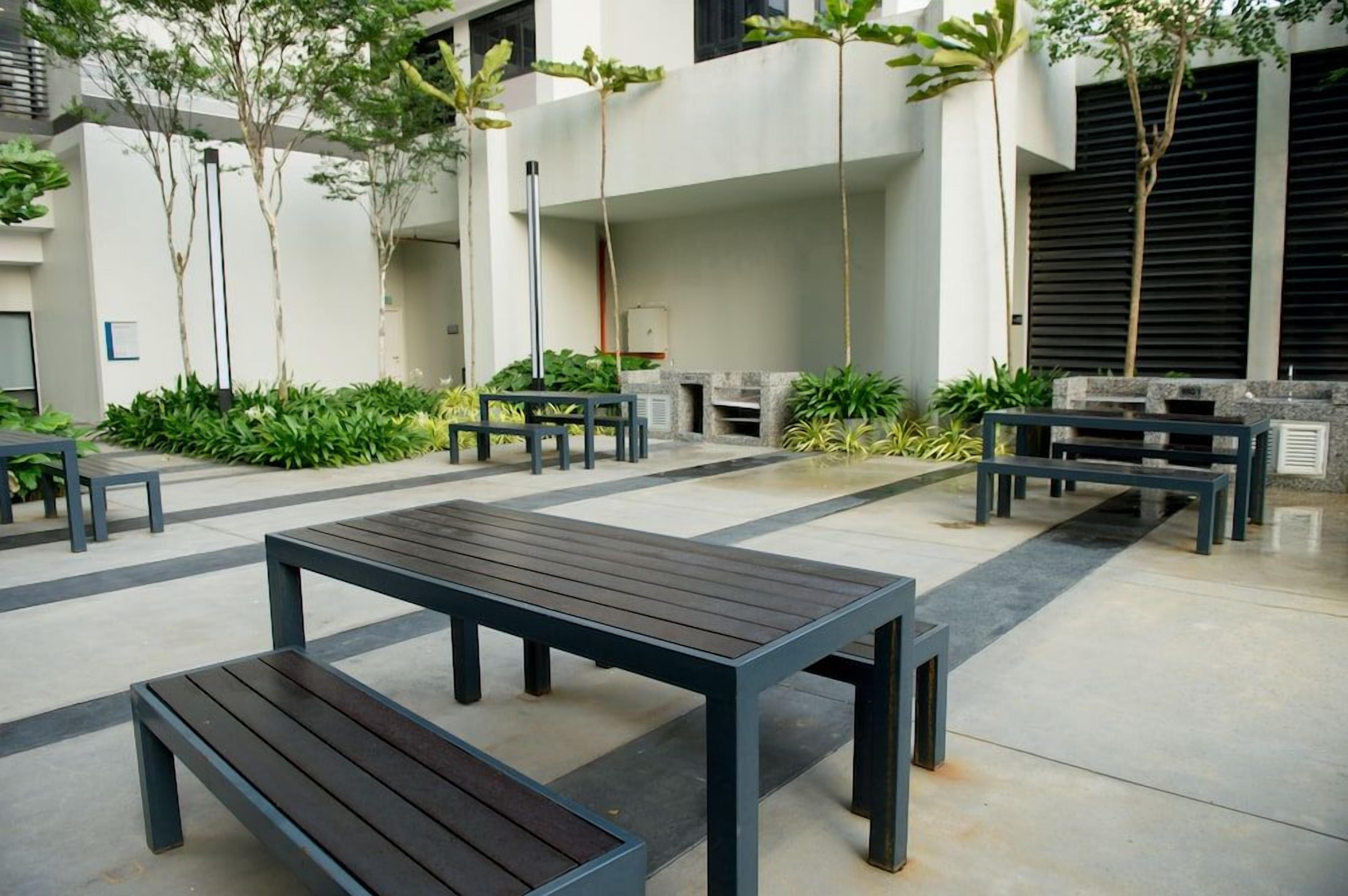 An outdoor seating area with tables and benches, surrounded by greenery and a modern building facade.