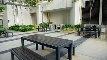 An outdoor seating area with tables and benches, surrounded by greenery and a modern building facade.