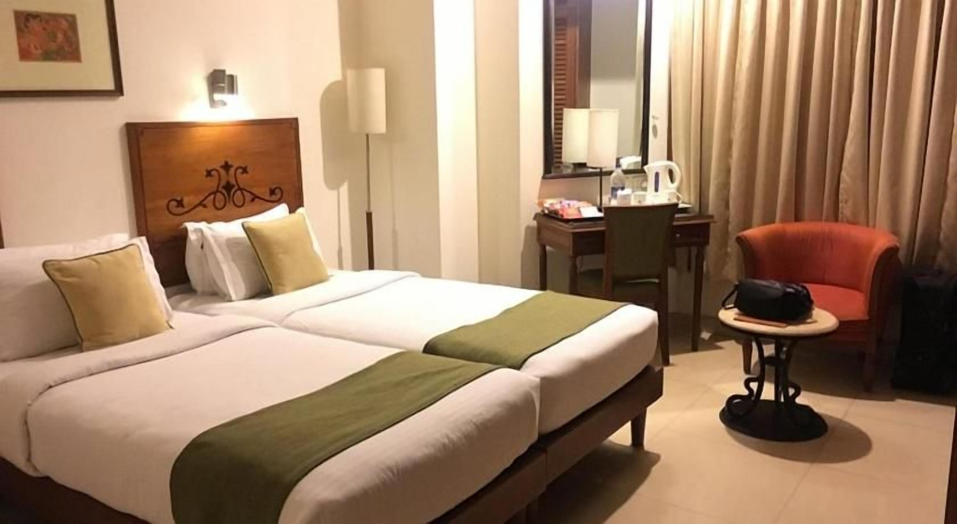 A hotel room with a double bed, a desk with a chair, a small table with a bag, and a red chair.