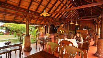 An indoor dining area with wooden tables and chairs, a thatched roof, and a view of greenery outside.