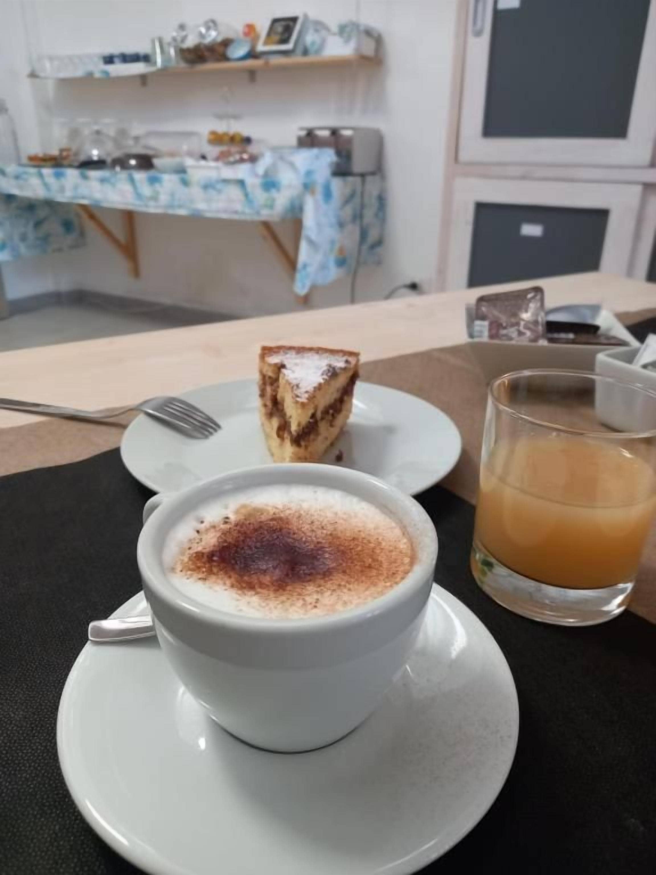 A cup of coffee with a slice of cake on a plate, a glass of orange juice, and a fork on a table.