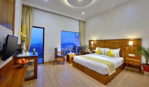 A hotel room with a bed, a desk with a television, a chair, a nightstand, a potted plant, and a view of the outdoors. - Chail Villas (Shimla)