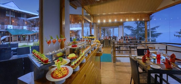 A buffet setup with various food items and drinks, a wooden floor, and a view of the outdoors. - Chail Villas (Shimla)