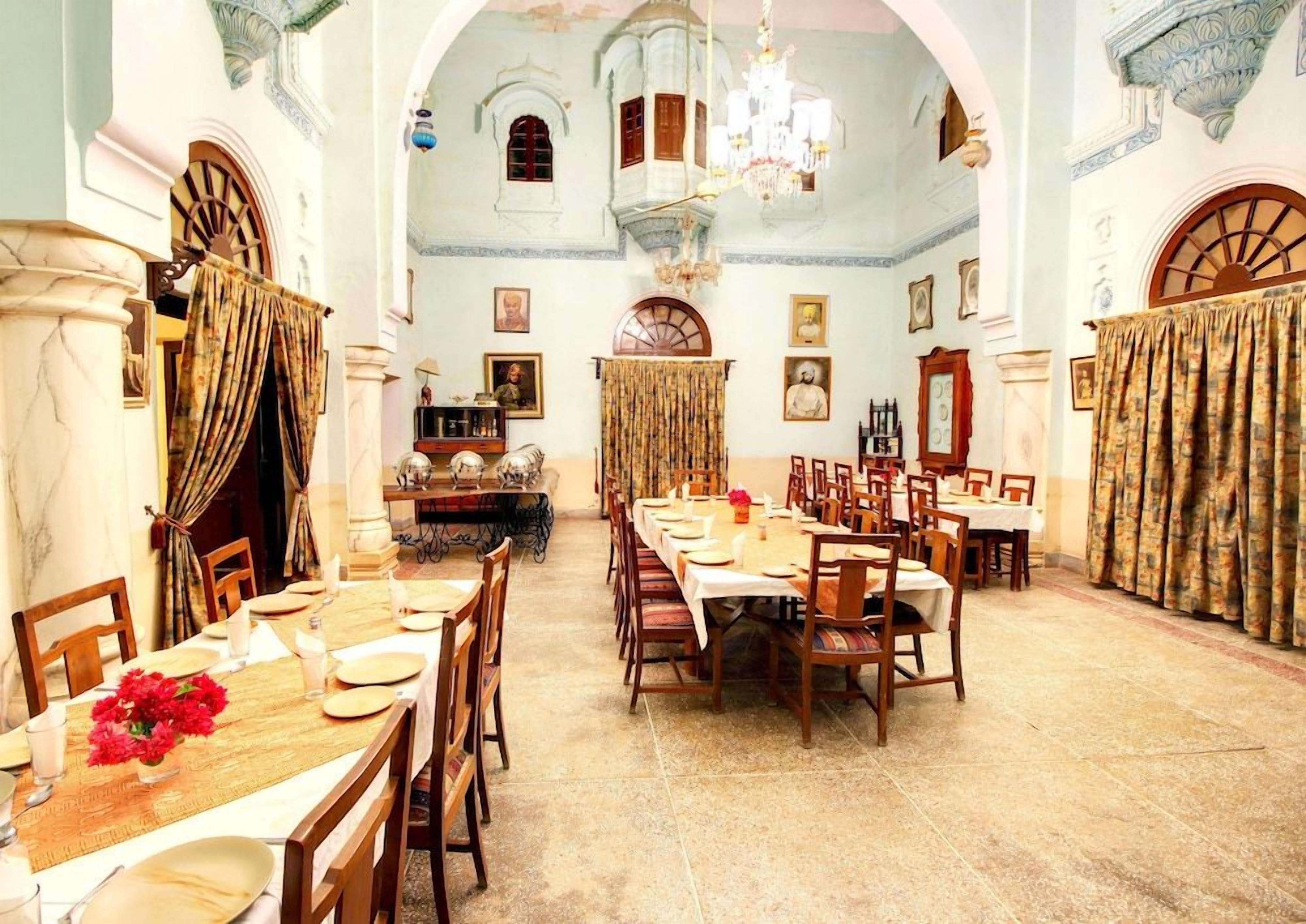 A dining hall with a long table set for a meal, surrounded by chairs, with a chandelier above.