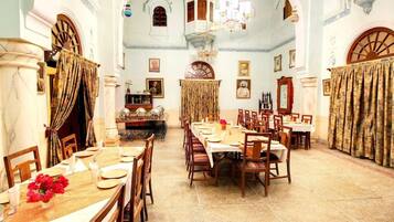 A dining hall with a long table set for a meal, surrounded by chairs, with a chandelier above.
