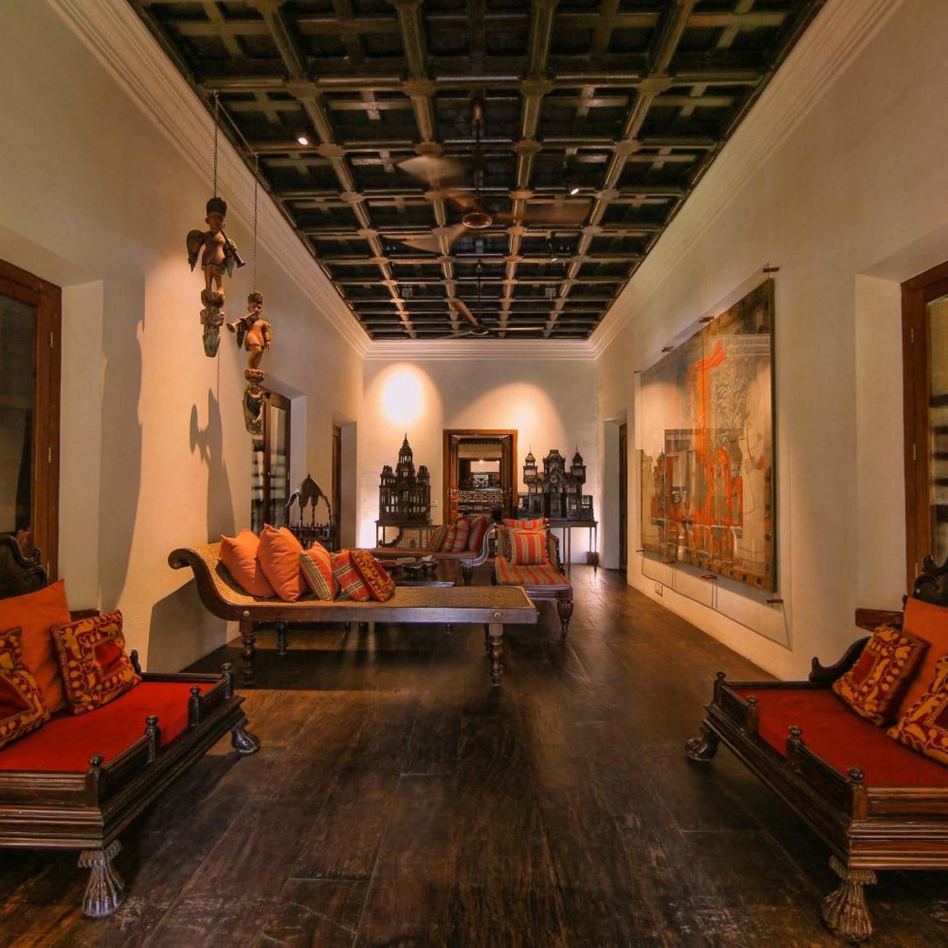 A room with a high ceiling, ornate ceiling design, a sofa set with patterned cushions, a table, and a large framed artwork on the wall.