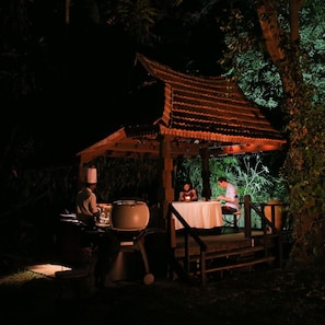 An outdoor dining area with a traditional pavilion structure, a barbecue grill, and a table set for two.