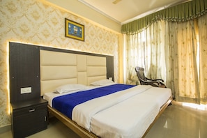 A hotel room with a large bed, a nightstand, a chair, and a window with curtains. - Hotel Jhanvi Dale (Haridwar)