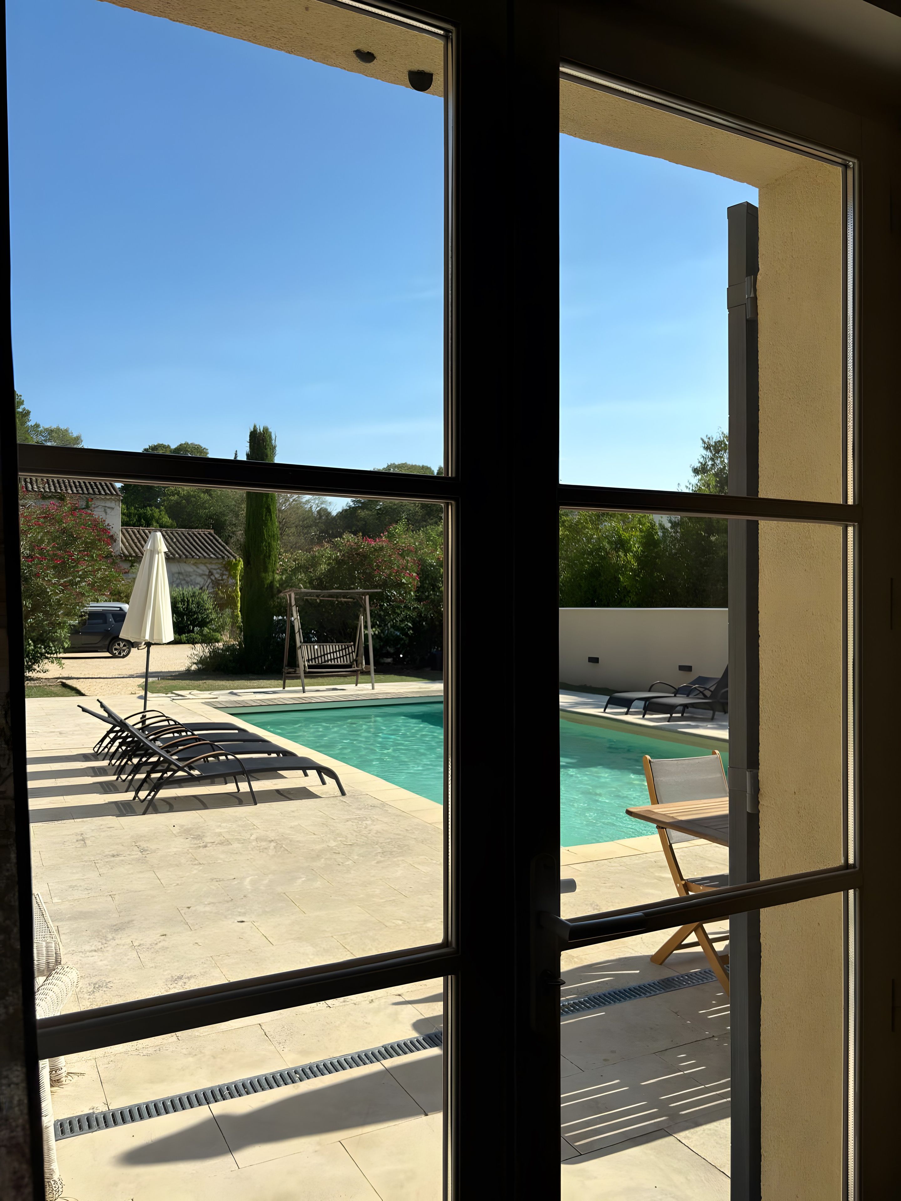 Blick durch ein Fenster auf einen Poolbereich mit Liegestühlen und einem Sonnenschirm.