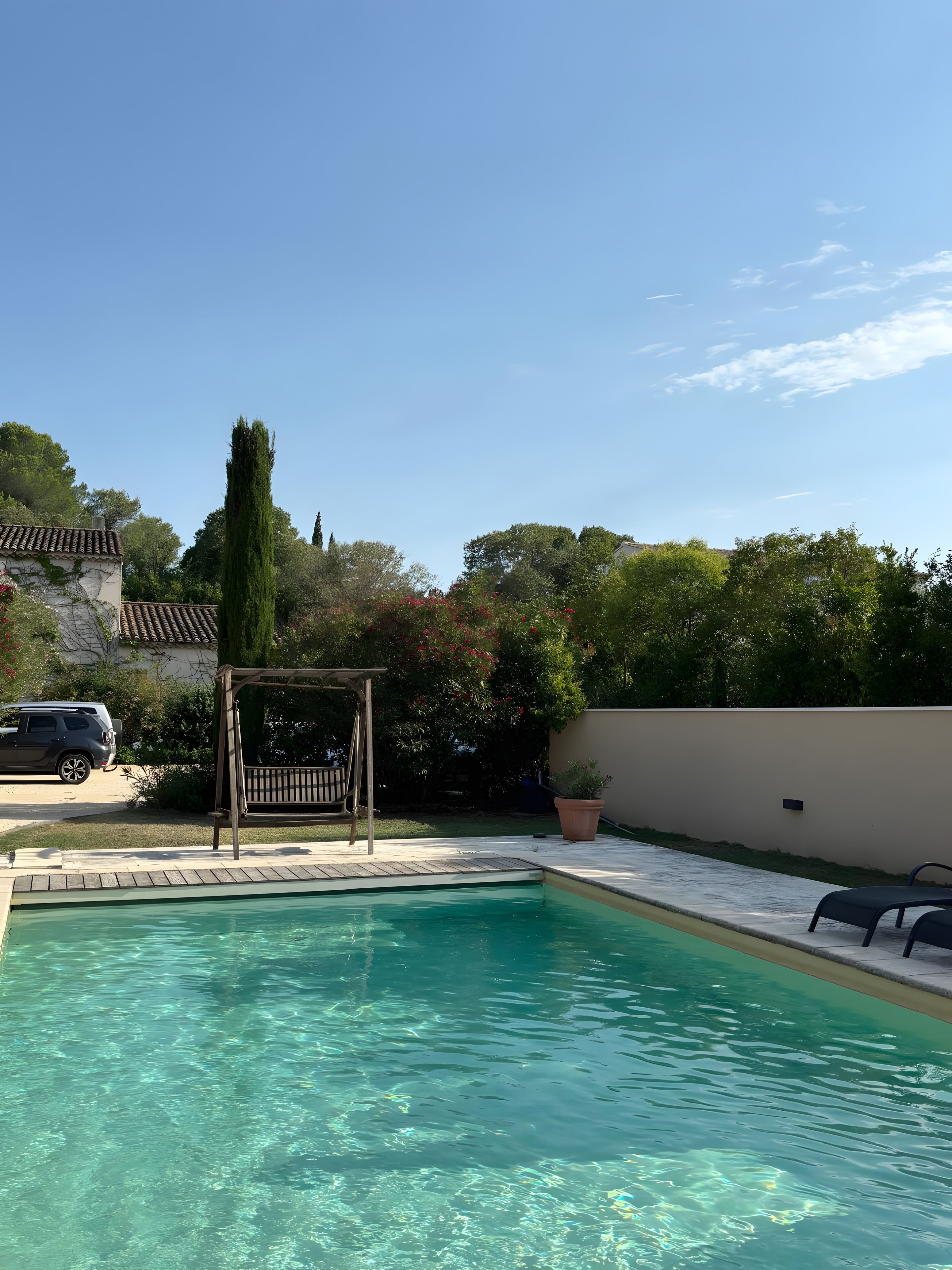 A clear swimming pool with a wooden deck, a pergola, and a car parked nearby.