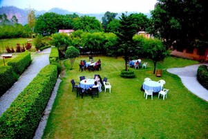 Un jardín al aire libre con varias mesas y sillas, rodeado de setos y árboles bien cuidados.