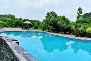 Piscina azul cristalina con un muro de piedra, rodeada de vegetación exuberante y una mesa con sillas bajo un paraguas rosa.