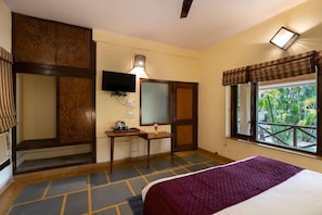A hotel room with a bed, a desk, a television, and a window with a view of greenery. - Corbett Wild Resort (Ramnagar)