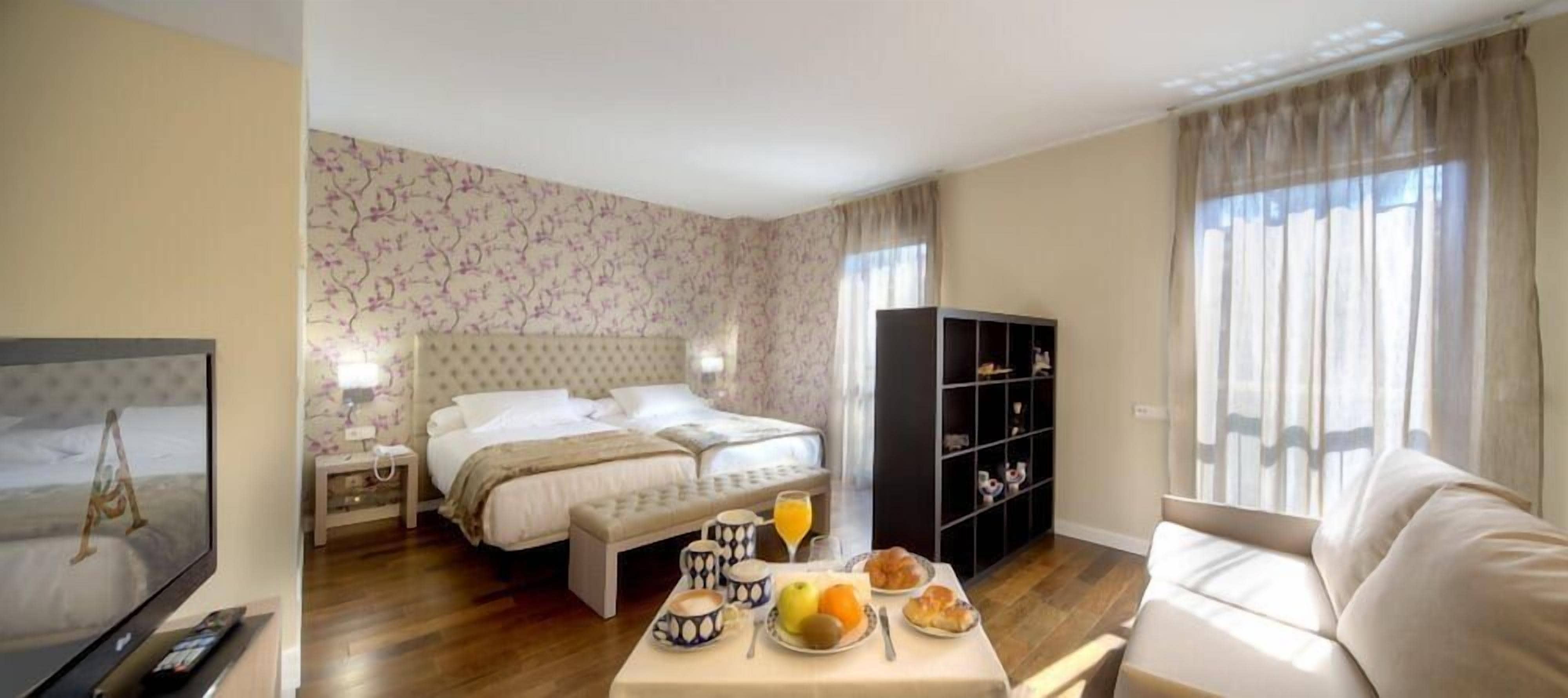 A hotel room with a bed, bedside table, a TV, a dining area with a table and chairs, and a bookshelf.