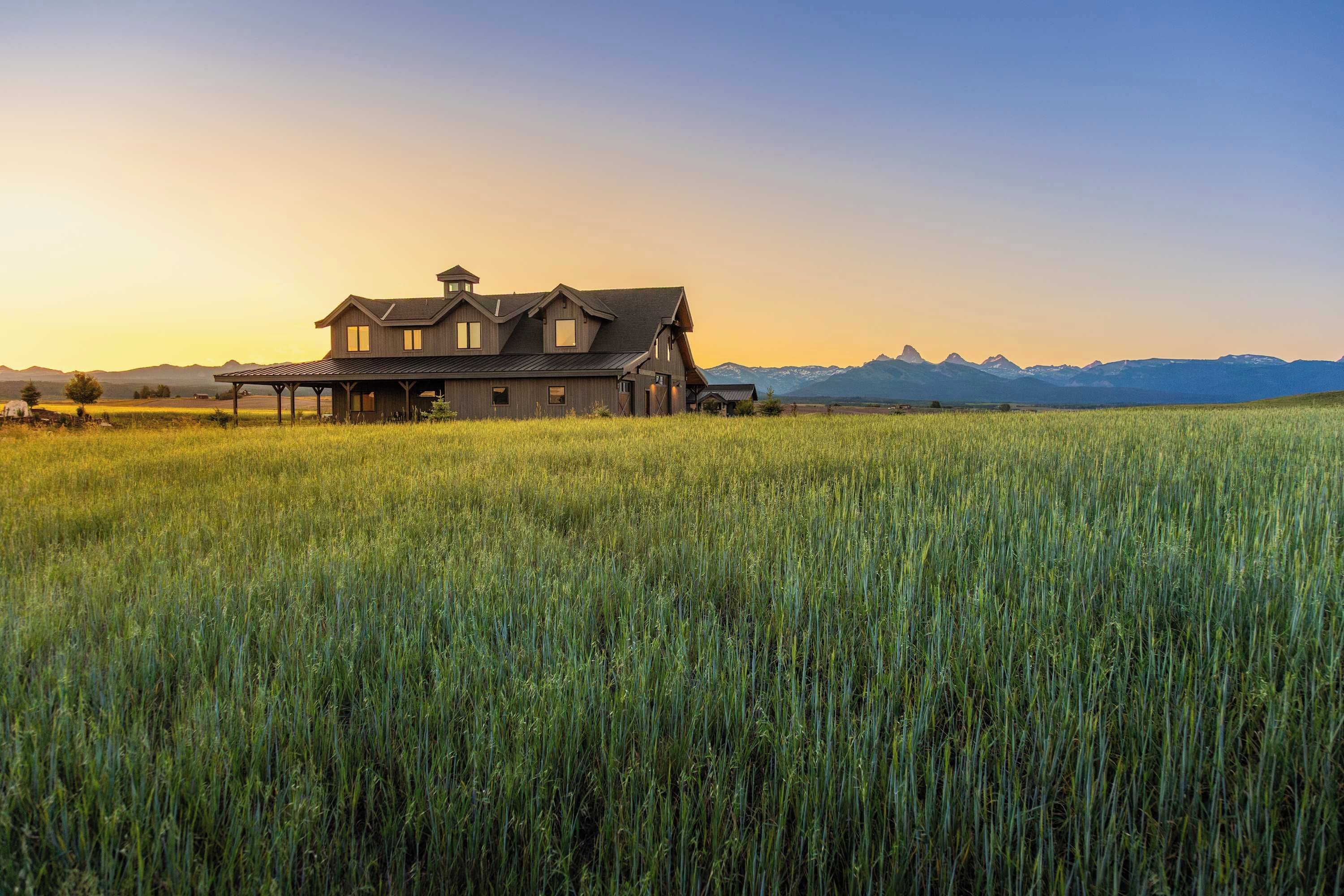A large house with a covered porch, surrounded by a field of tall grass, with mountains in the background.