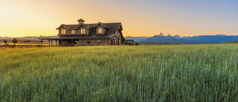 Une grande maison dotée d’un porche abrité, entourée d’un champ d’herbes hautes, avec des montagnes en arrière-plan.