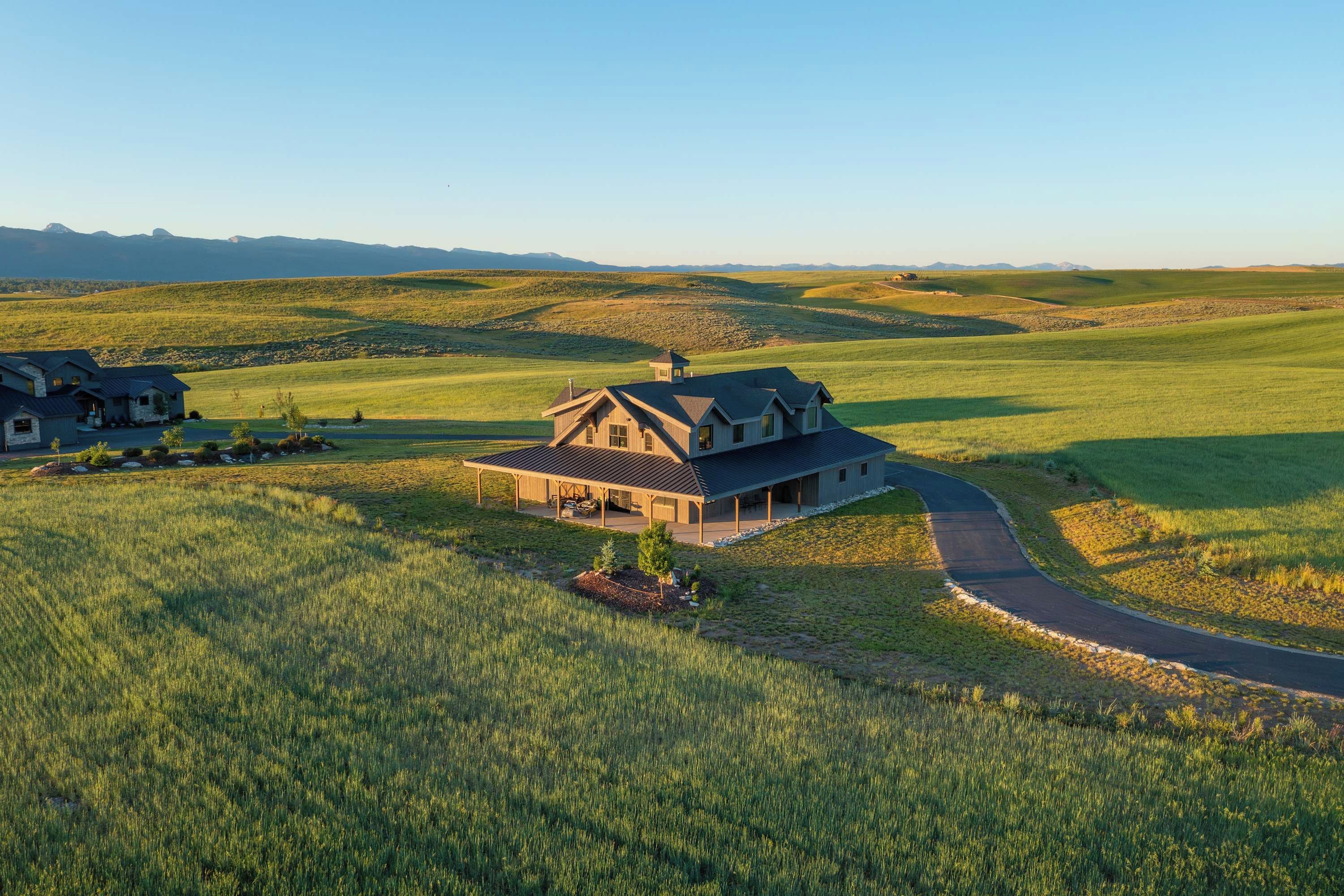 A large house with a covered porch, surrounded by expansive green fields and a winding road.