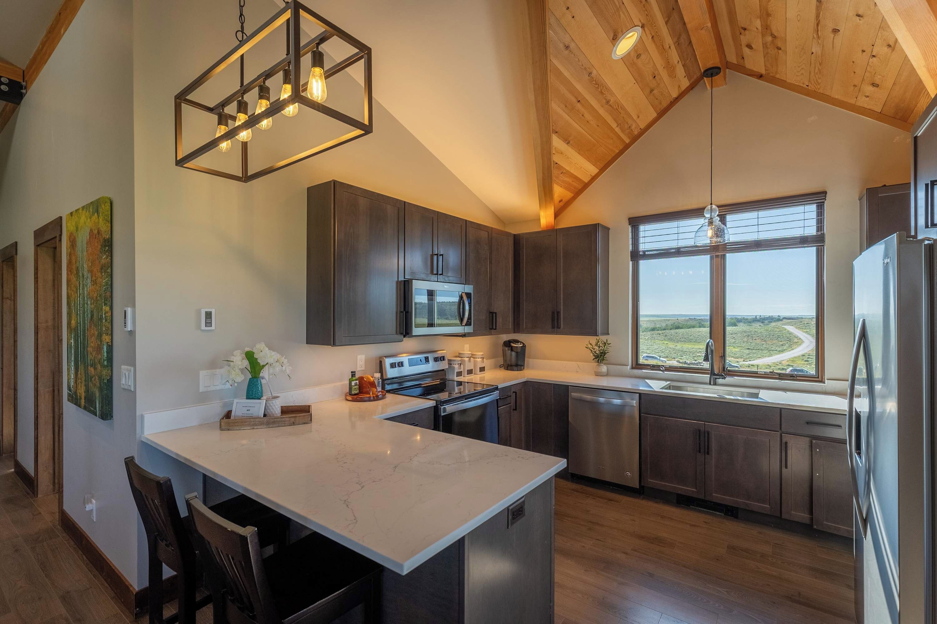 A modern kitchen with dark wood cabinets, a central island, stainless steel appliances, and a large window offering a view of the landscape.