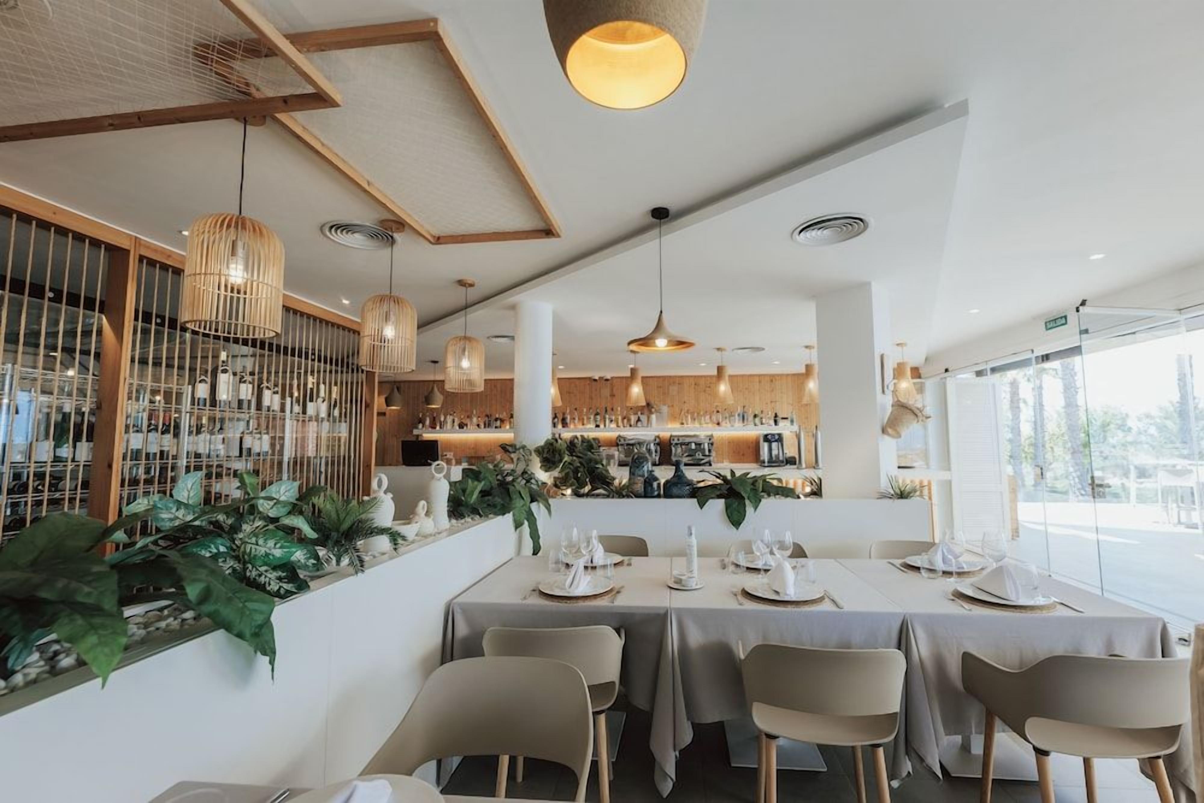Un interior de restaurante con mesas dispuestas para comer, lámparas colgantes y una zona de bar al fondo.