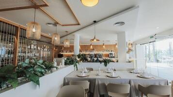 Un interior de restaurante con mesas dispuestas para comer, lámparas colgantes y una zona de bar al fondo.