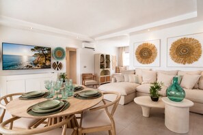 A modern living room with a dining area, featuring a round wooden table set for four, a beige sofa, and decorative wall art.
