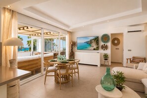 A modern living room with a dining area, large windows with a view of the sea, and a flat-screen TV mounted on the wall.