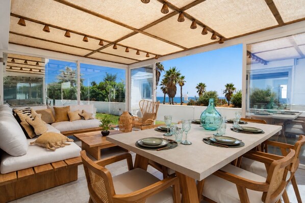 A sunlit dining area with a view of the sea, featuring a table set with plates, glasses, and a decorative vase.