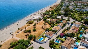 An aerial view of a coastal residential area with a beach, swimming pools, and houses with tiled roofs.