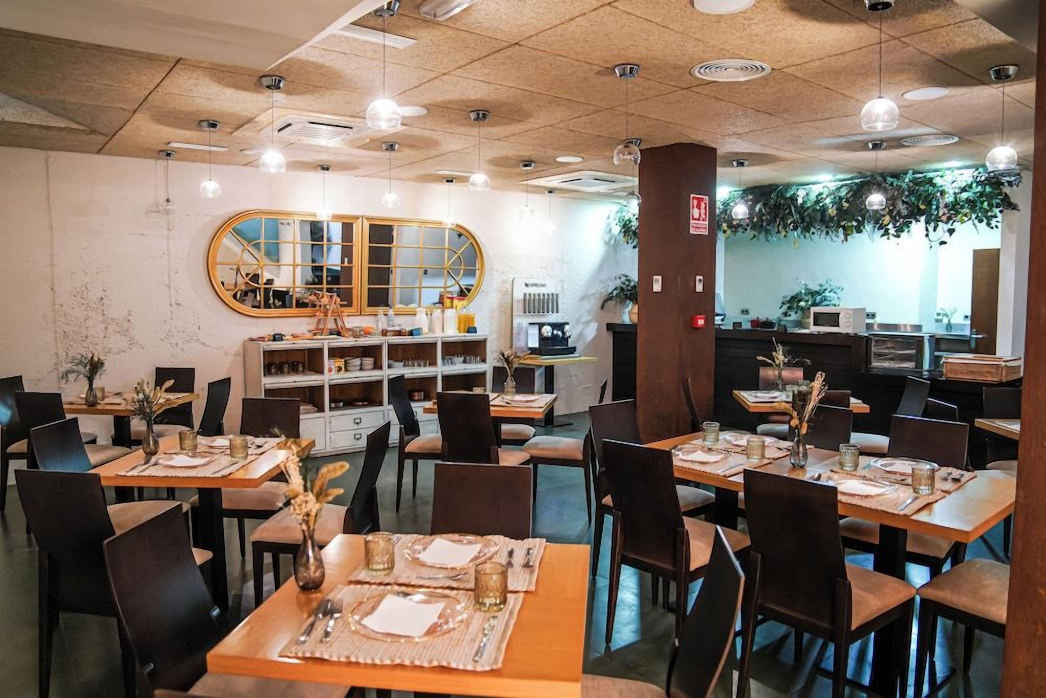 Interior de restaurante con varias mesas, sillas, lámparas colgantes, un espejo grande y una zona de cocina visible al fondo.