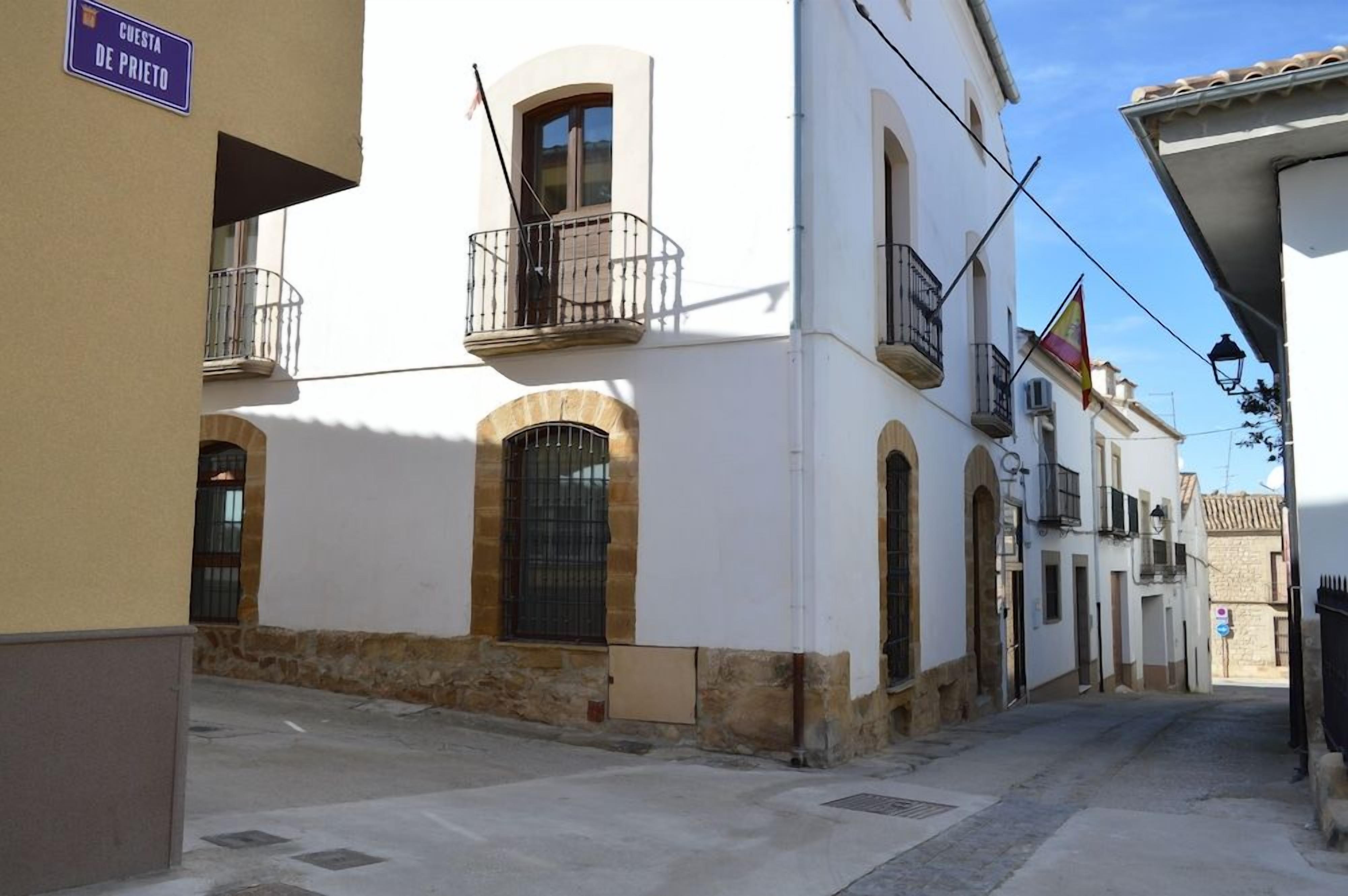 Una calle estrecha con edificios blancos, un cartel que dice 'CUESTA DE PRIETO' y una bandera.