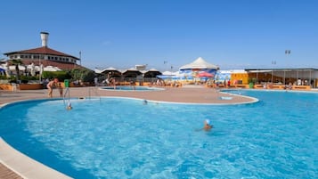 En stor udendørs swimmingpool med flere baner, omgivet af et overdækket opholdsområde og en bygning med en markant skorsten.