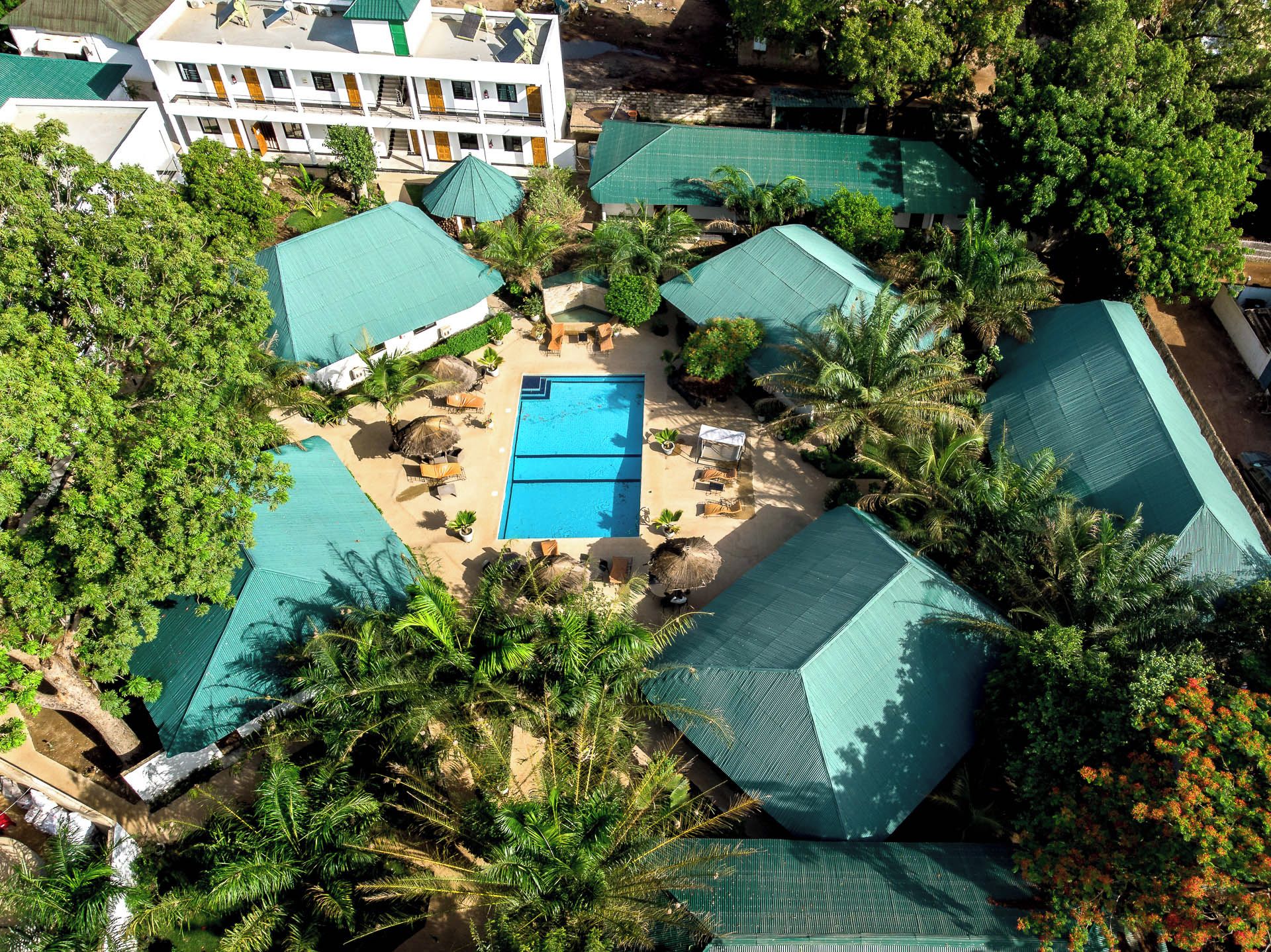 An aerial view of a resort with a central swimming pool surrounded by palm trees and covered structures.